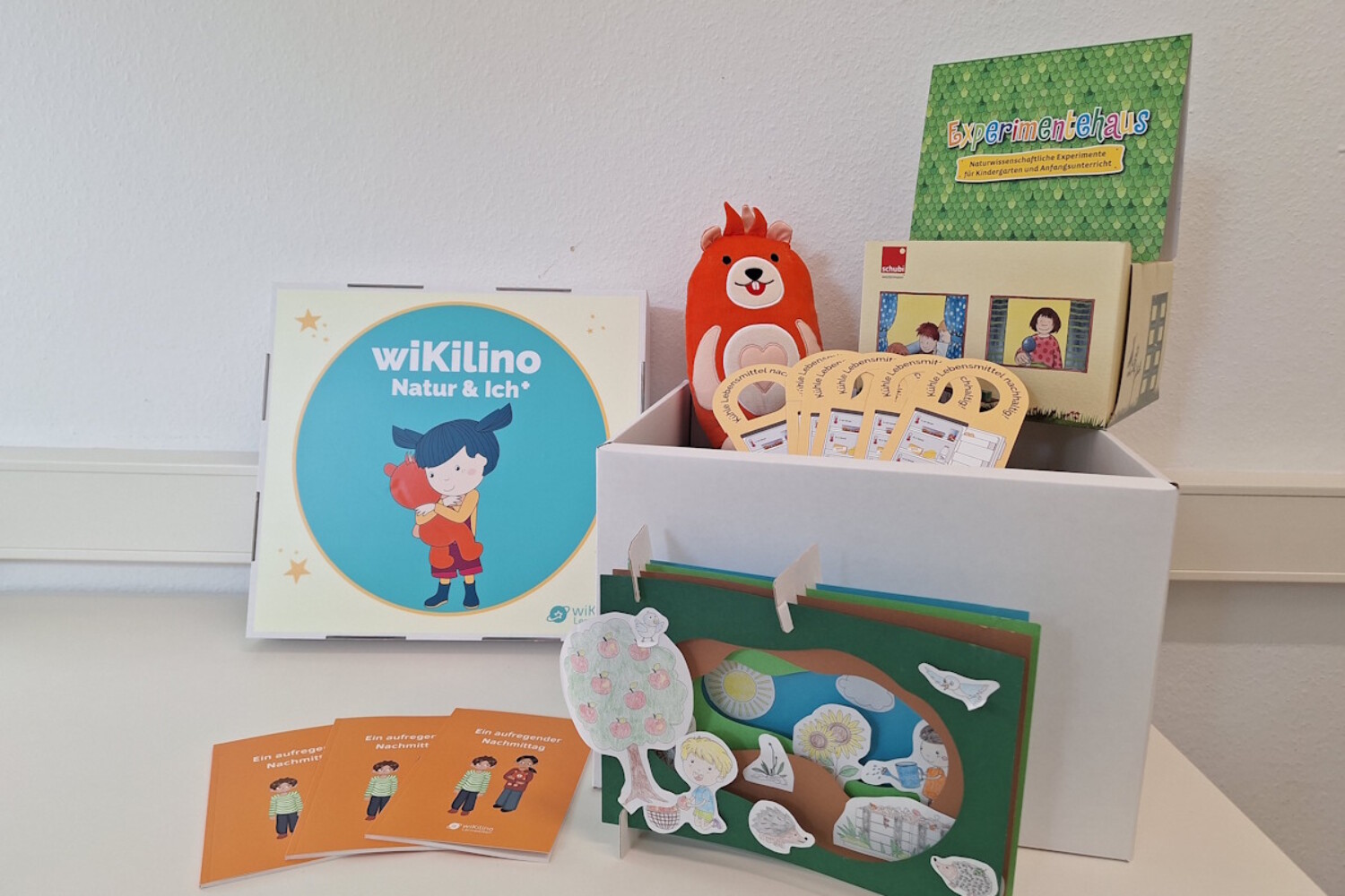 Mehrere wiKilino-Materialien zu Natur & Ich und dem Experimentehaus liegen vor einer Wand, mit Karten, Heften und Stofftier.