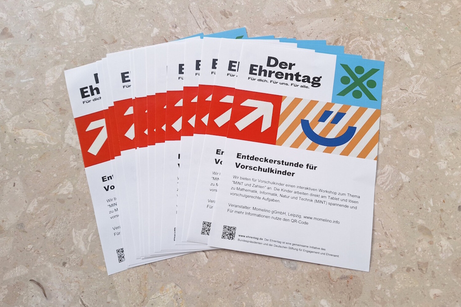 Mehrere Flyer zum Ehrentag liegen fächerförmig auf einem Steinboden und zeigen „Entdeckerstunde für Vorschulkinder“.