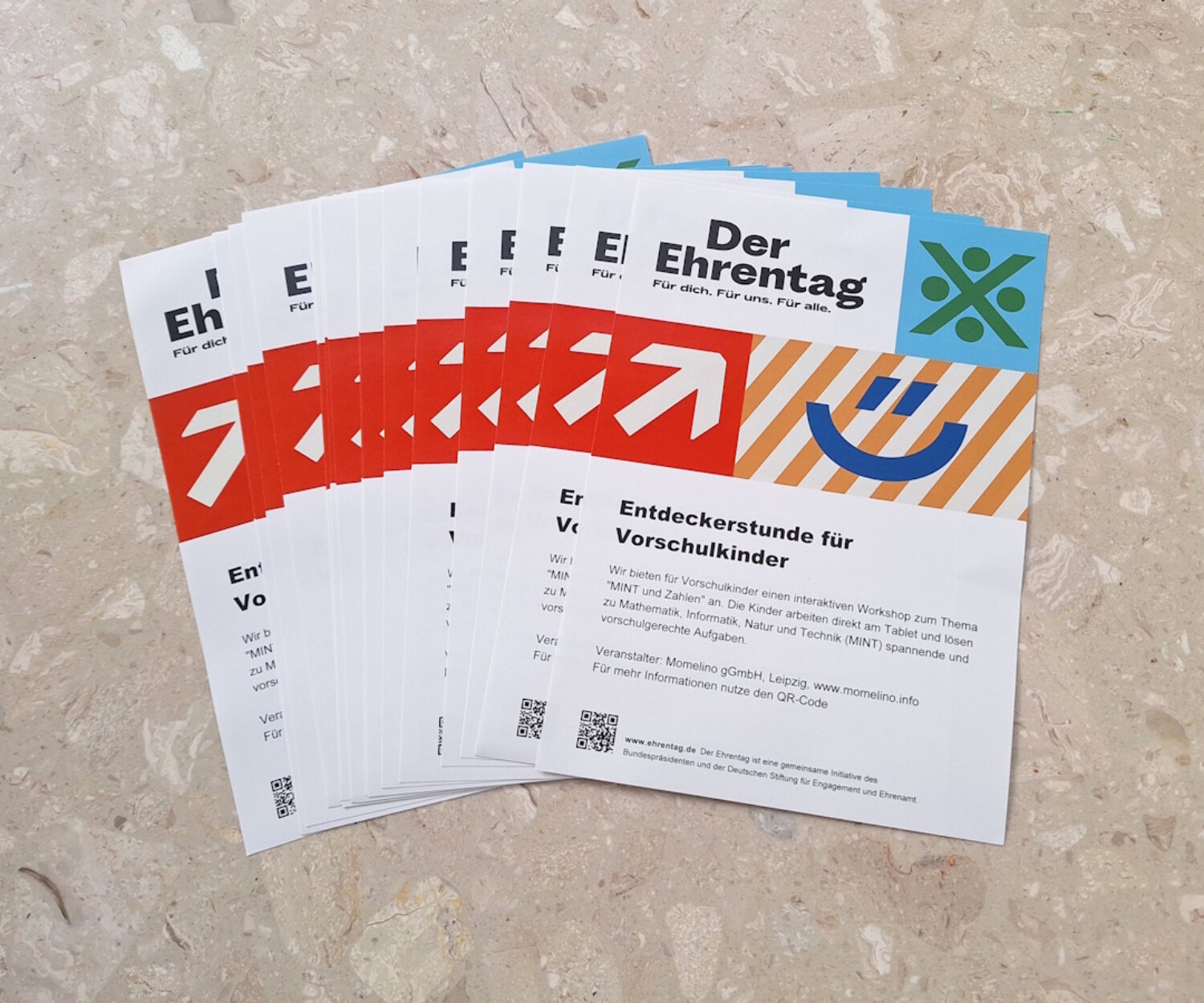 Mehrere Flyer zum Ehrentag liegen fächerförmig auf einem Steinboden und zeigen „Entdeckerstunde für Vorschulkinder“.