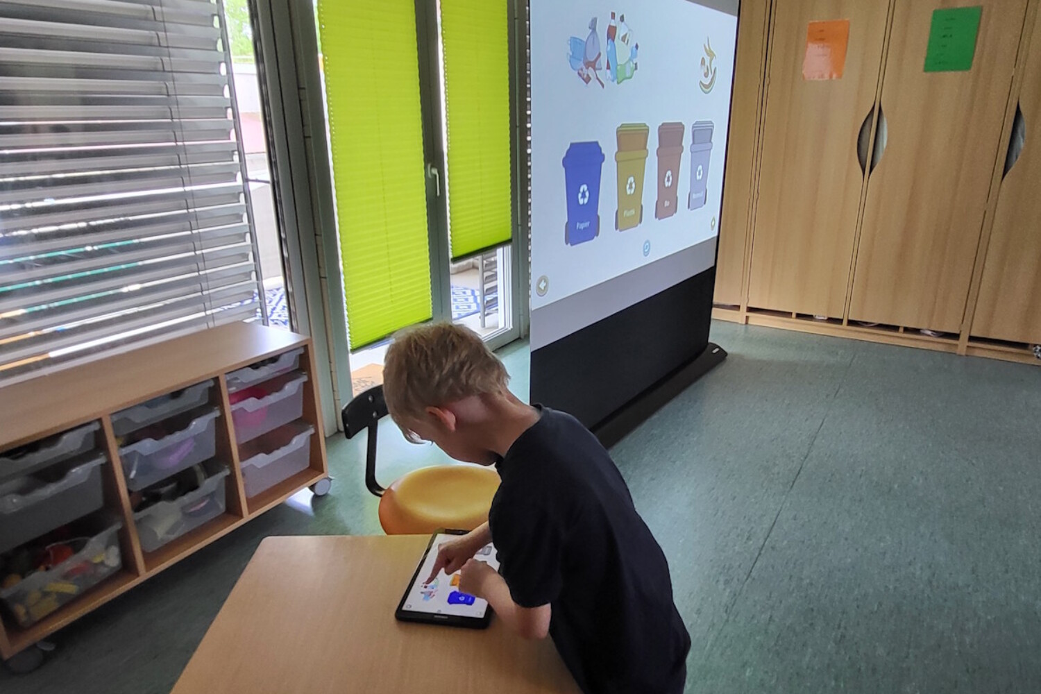 Ein Junge tippt an einem Tablet, während auf der Leinwand im Klassenzimmer Mülltrennung mit farbigen Tonnen gezeigt wird.