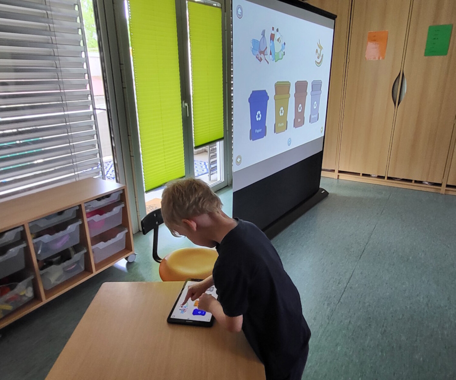 Ein Junge tippt an einem Tablet, während auf der Leinwand im Klassenzimmer Mülltrennung mit farbigen Tonnen gezeigt wird.